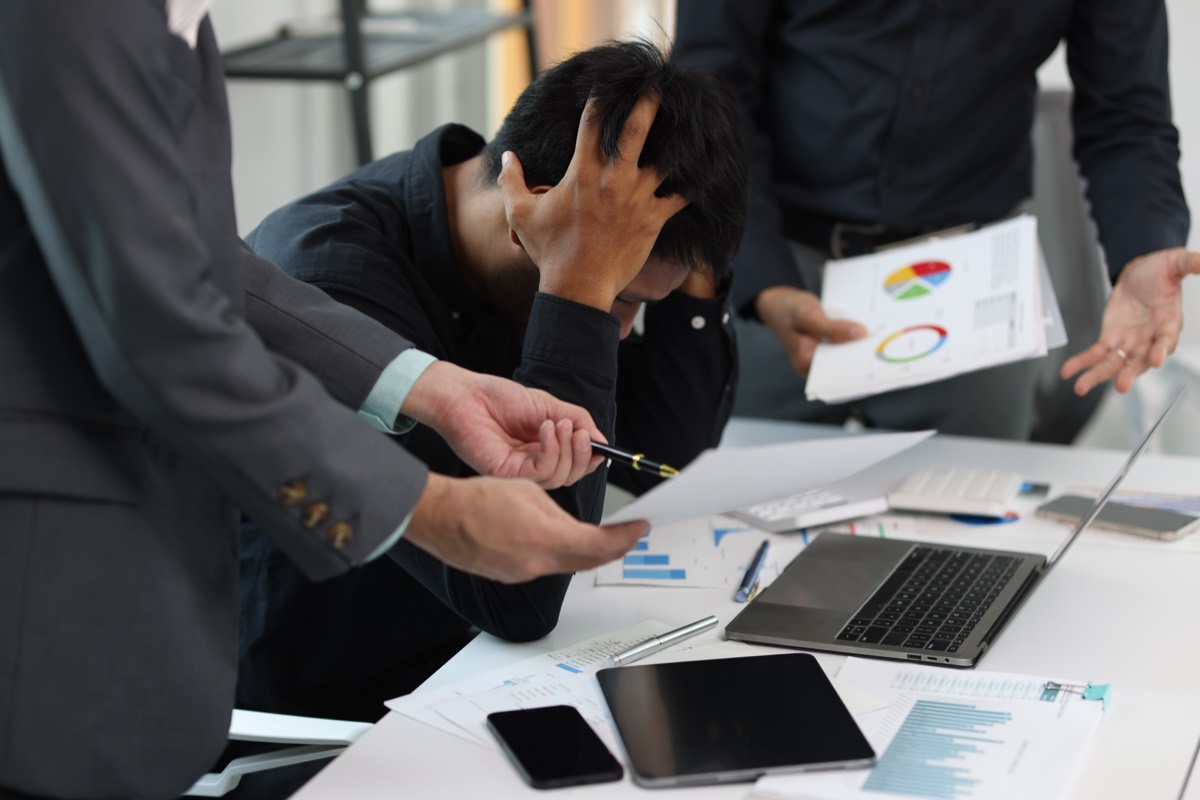 An overwhelmed employee holds his head while colleagues push more paperwork at him
